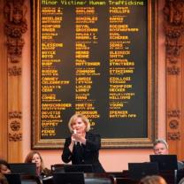 Blonde woman speaking to a crowd in front of a sign with names of people who were victims of human trafficking