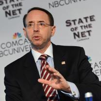 Balding white man with wire rimmed glasses wearing a suit speaking and make a hand gesture