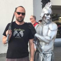 Guy with dark beard, sunglasses and sword next to a silver statue of a half-naked warrior with metal helmet