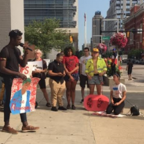 Young black man speaking into a mic to a crowd of people outside with signs