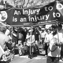 Black and white photo of big banner with words An Injury to one is an injury to all and mostly black people marching with it, some with drums