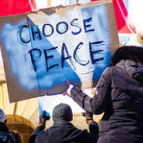 Woman with her back to the camera wearing a large winter coat with furry hood holding a big sign that reads CHOOSE PEACE, she is with a group of people at a rally