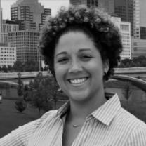 Smiling black woman with short cropped curly hair and a striped button down shirt standing with downtown buildings and a bridge across the river behind her
