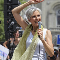 Jill Stein with her arm above her head and in a white tank top and yellow scarf outside at rally