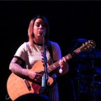 Young white woman in a white t-shirt playing a guitar and singing at a mic with a black background