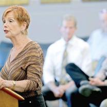 Middle-aged woman with red hair standing at a podium talking and people behind her in audience