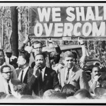 Black and white photo of Martin Luther King Jr. and others marching under sign that says We Shall Overcome