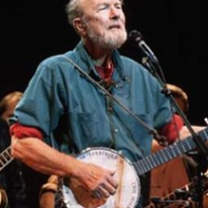 Older gray haired man with beard playing a banjo and singing into a mic