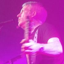 Purple lighted picture of white man performing side view holding a guitar