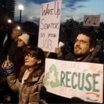 People standing outside in winter coats at dusk one white man with brown hair, facial hair and glasses with a sign that reads Recuse and a white woman next to him 