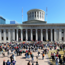 Big government building with round tower at top and white columns and lots of people on the grounds in front