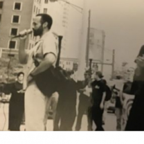 Black and white grainy photo of black man speaking at a mic and people protesting on a street