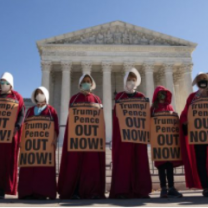 Proestors dressed like handmaidens outside a government building with signs saying Trump Pence Out Now