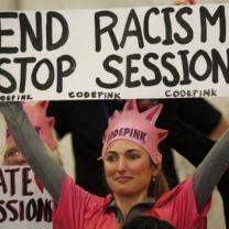 Blonde woman in pink with Code Pink crown holding a sign that says End racism Stop Sessions