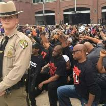 Lots of black men down on one knee with fists raised in the street with police man in foreground