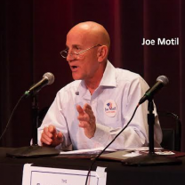 Bald white man with little wire-rimmed glasses talking at a mic on a table, wearing a white shirt and gesturing with his left hand