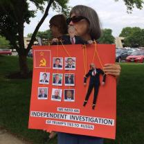 Woman standing outside with sunglasses on holding a huge red sign in front of her asking for an independent investigation of Trump's ties to Russia