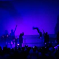 Silhouettes of people dancing and flipping in the air against a bright blue background on a stage in front of an audience