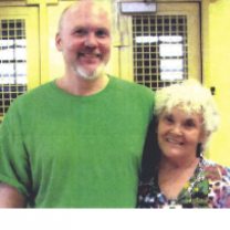 Tall bald white man with a mustache and beard and green T-shirt standing smiling with his arm around a gray haired smiling lady in a flowered dress