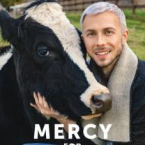 A man with white hair and goatee standing outside petting a black cow who has a white stripe up her nose and the word Mercy below