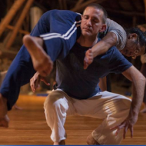White middle aged man squatting with his eyes closed holding a woman across is back in a dance move