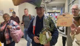 Man walking through airport holding a plant with people and balloons surrounding him